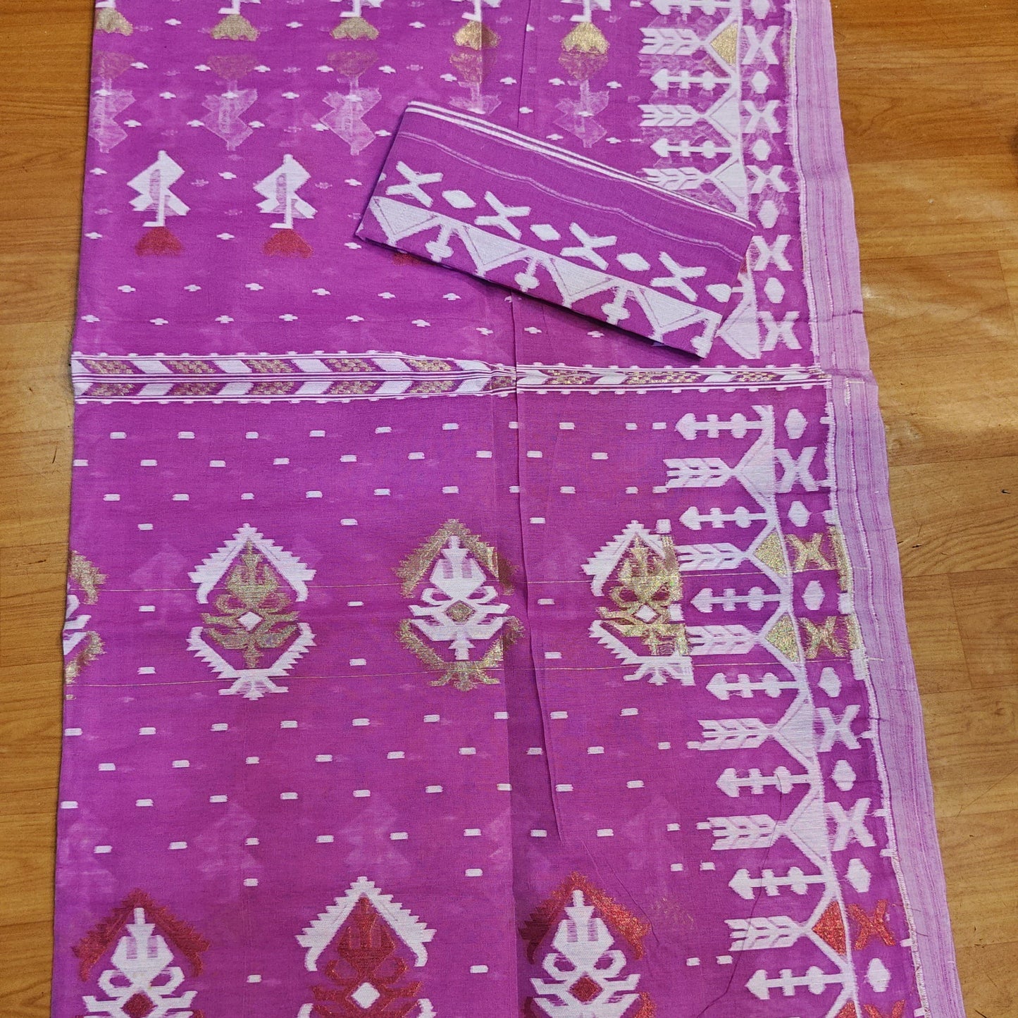 A purple and white patterned silk saree laid flat on a wooden floor, featuring traditional motifs and a matching fabric piece.