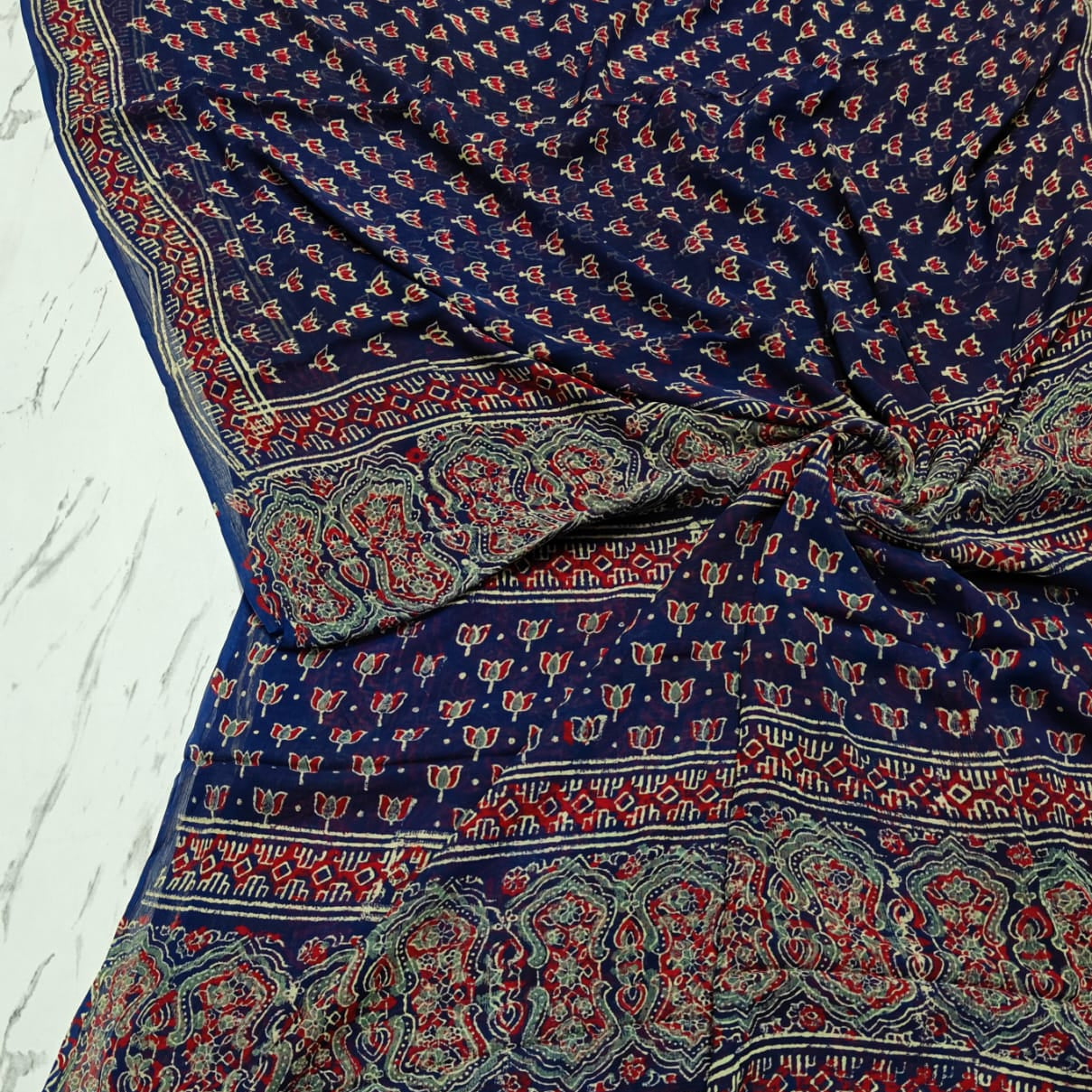 A navy blue chiffon saree with intricate red, beige, and white block-printed patterns and borders, draped on a light marble surface.