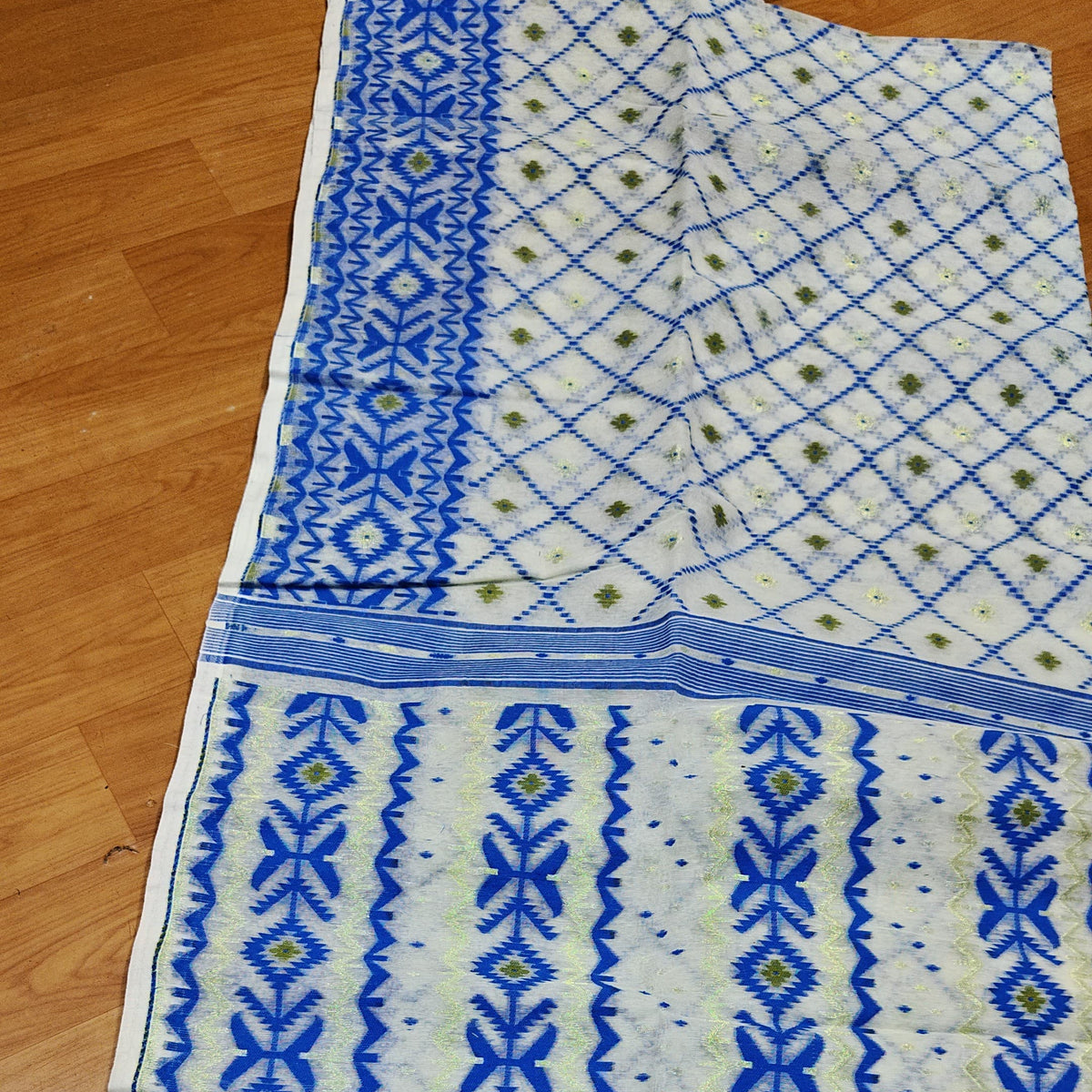 A cream-colored fabric saree or scarf with intricate blue and green geometric patterns, laid flat on a wooden floor.