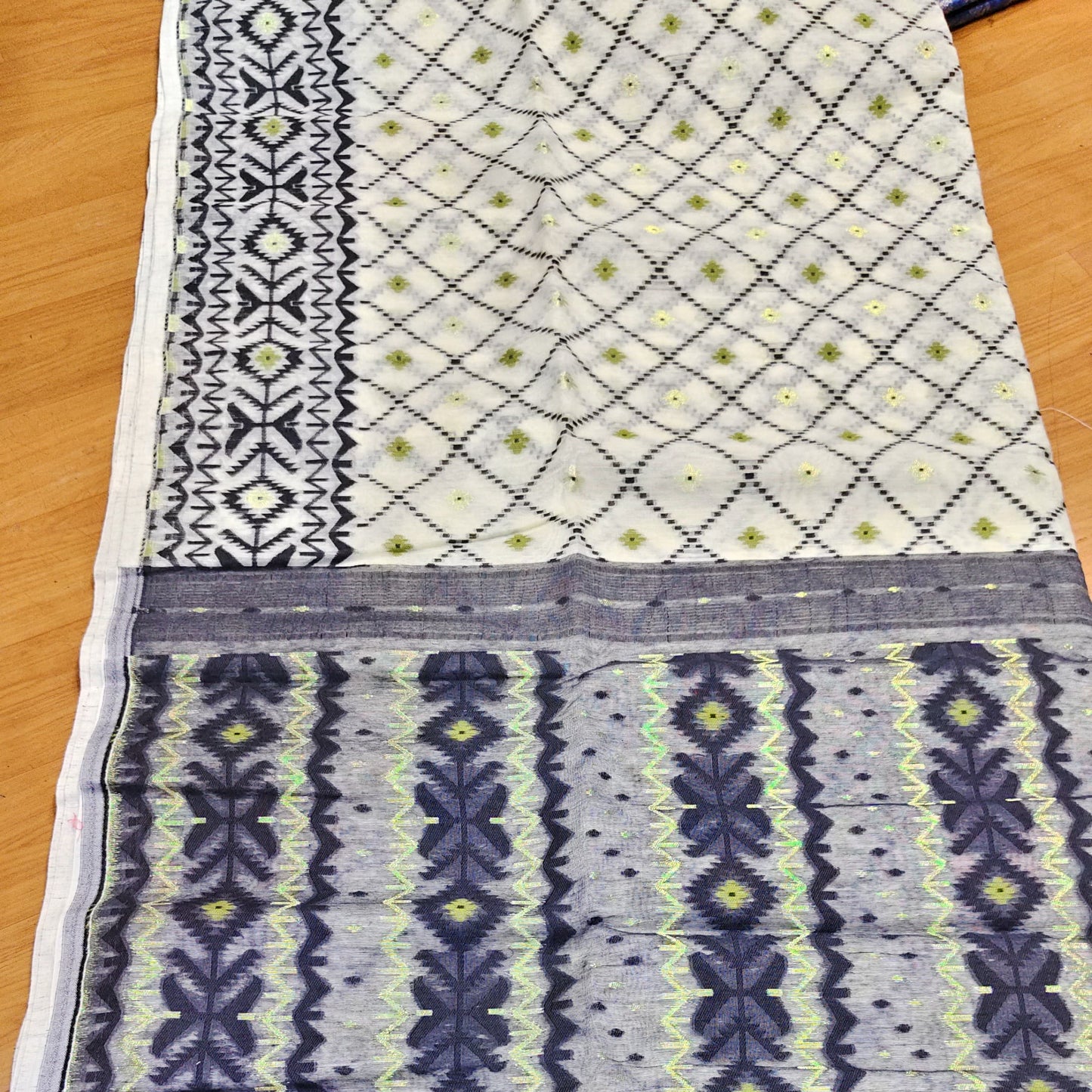A close-up of a patterned fabric, likely a saree or textile, laid flat on a wooden surface. The fabric features a cream base with black geometric borders, a black diamond lattice pattern with lime green dots, and vertical columns of dark blue-gray and black motifs with lime green accents.
