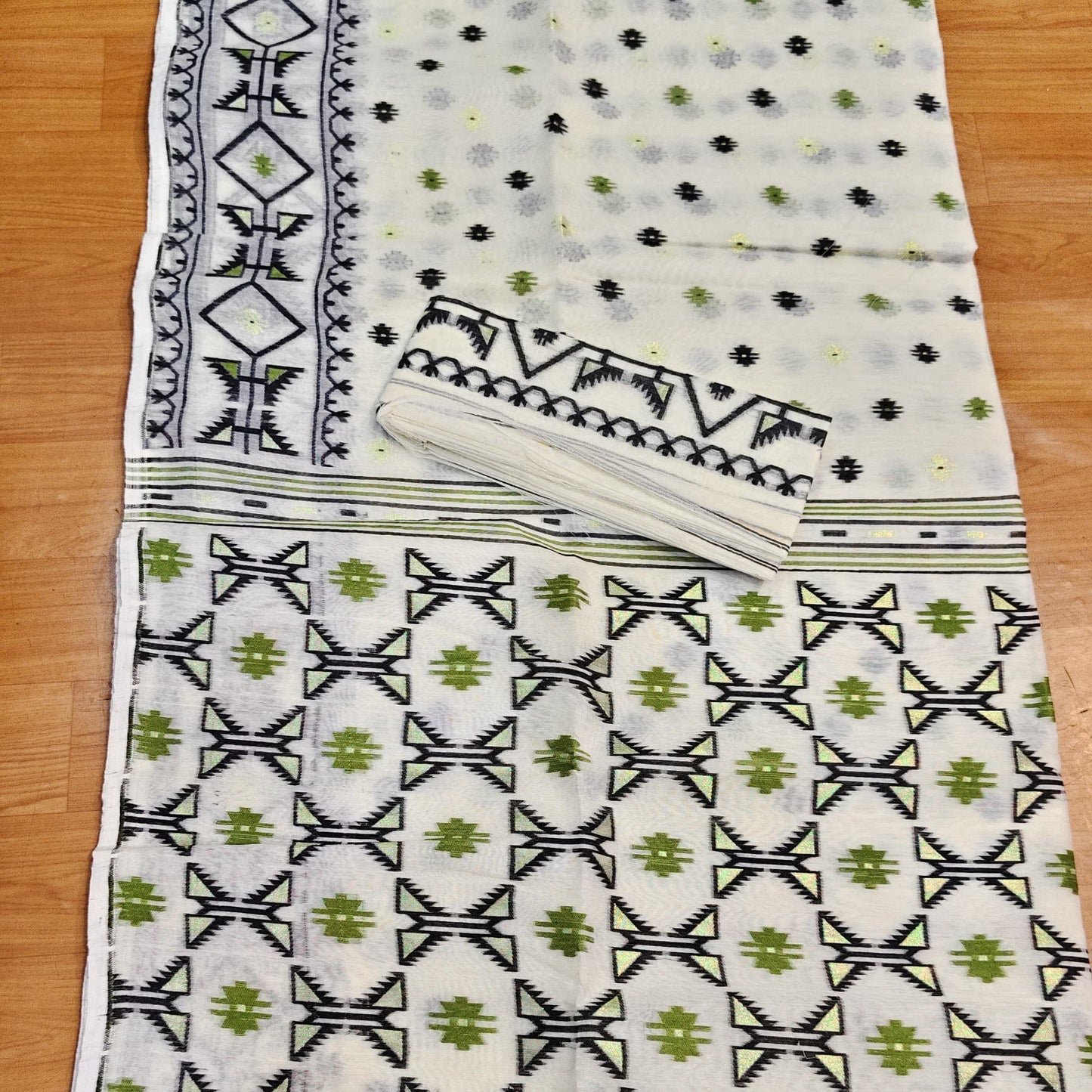 A white cotton saree with black and green geometric patterns, laid flat on a wooden floor. The fabric features a solid patterned border, a dotted body, and a wide border section with repeating bow and floral motifs.