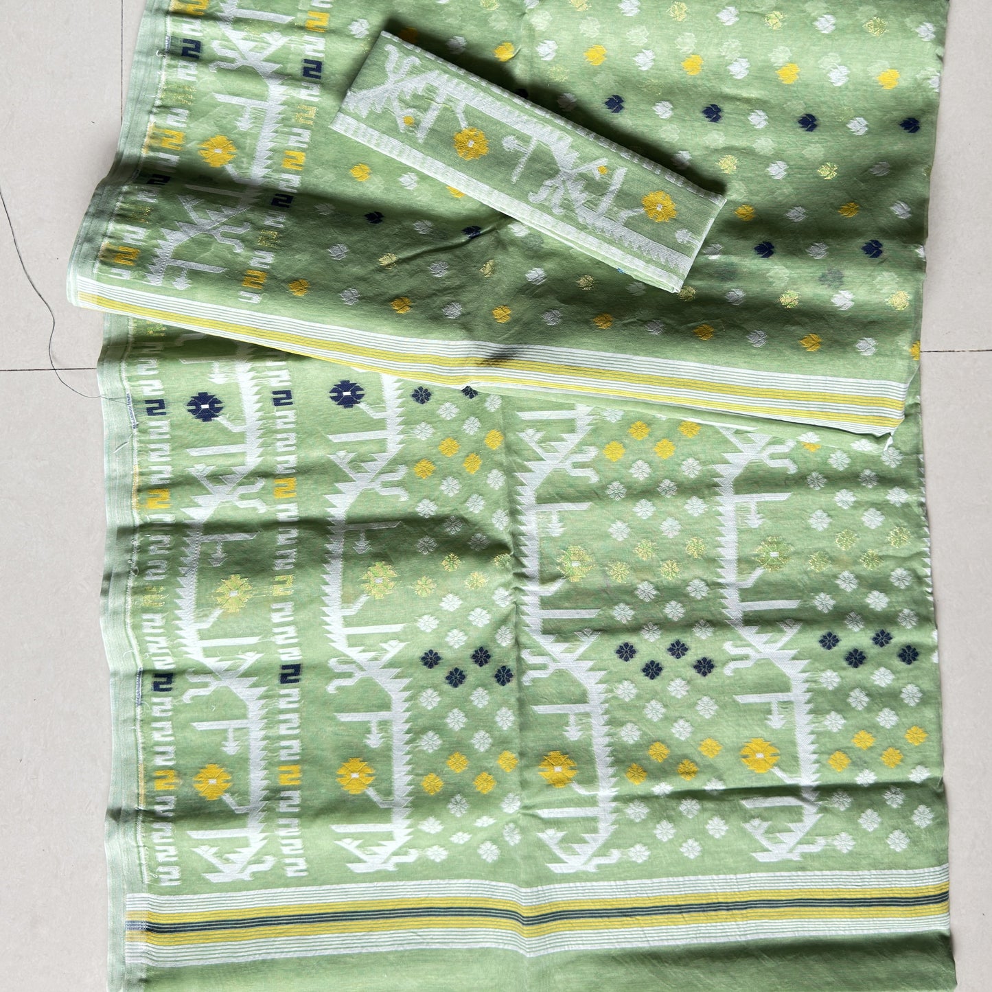 A folded light green fabric set with white and yellow printed patterns, laid flat on a light tiled floor. The set includes a main textile, a matching rectangular piece, and a smaller matching piece.