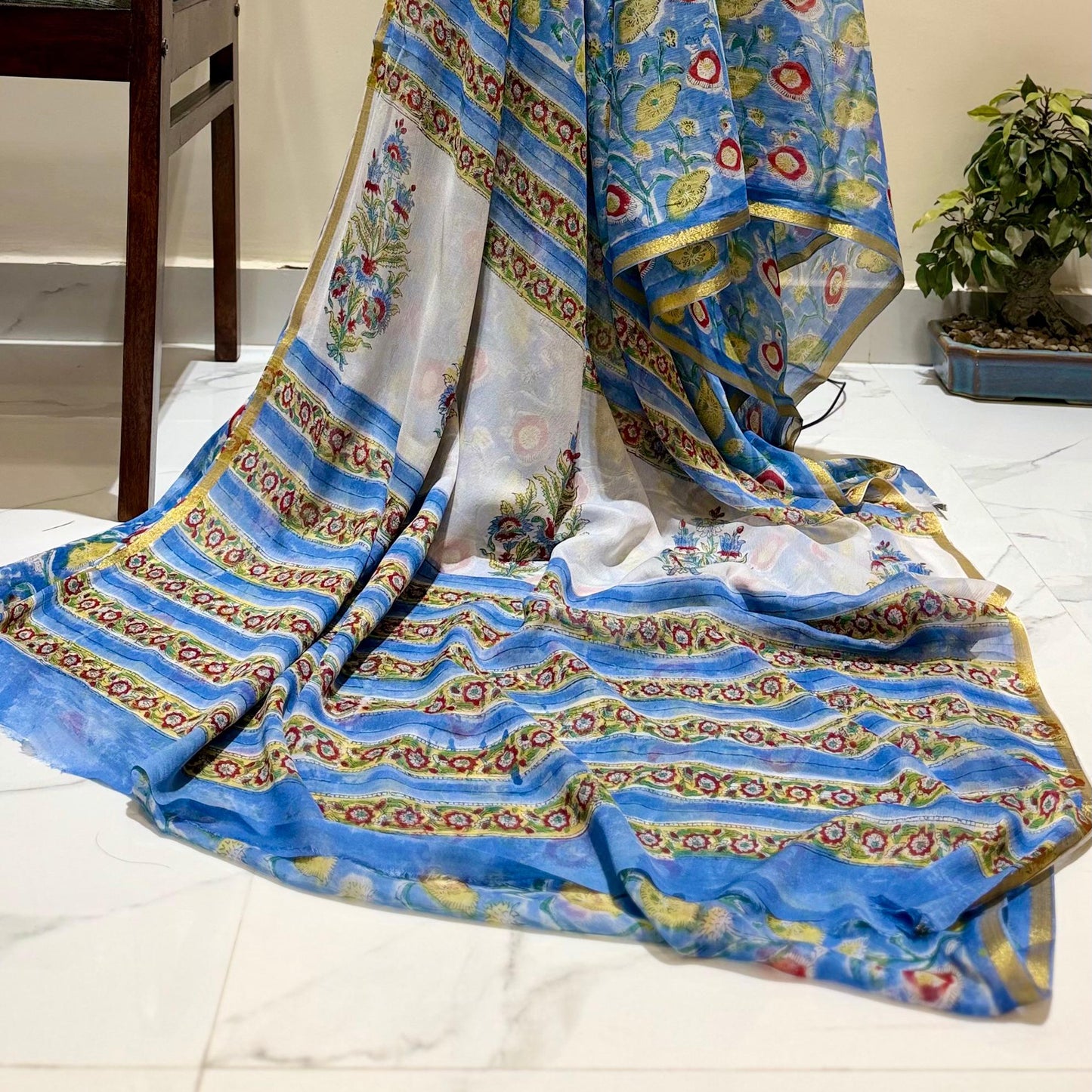 A blue patterned dupatta or scarf with intricate gold, red, and white motifs, draped over a chair and floor in an indoor setting.