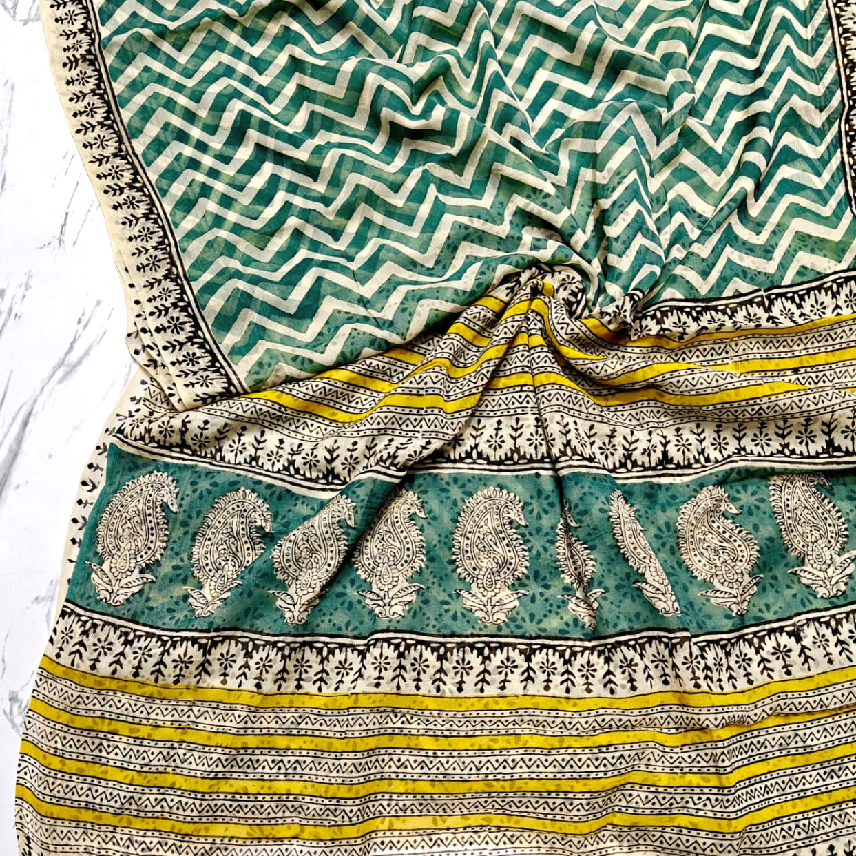 A folded chiffon saree with a green and white chevron pattern, a wide border with beige and black paisley motifs, and yellow and black stripes, displayed on a white marble surface.