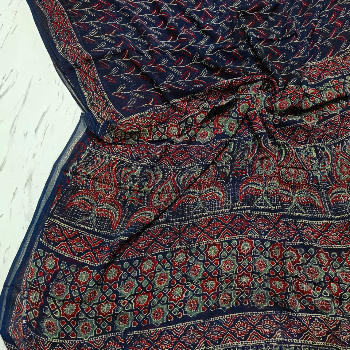 A navy blue block-print cotton saree with intricate red, green, and gold patterns, laid out on a light gray textured surface. A white label in the bottom-left corner reads 'chiffon saree'.