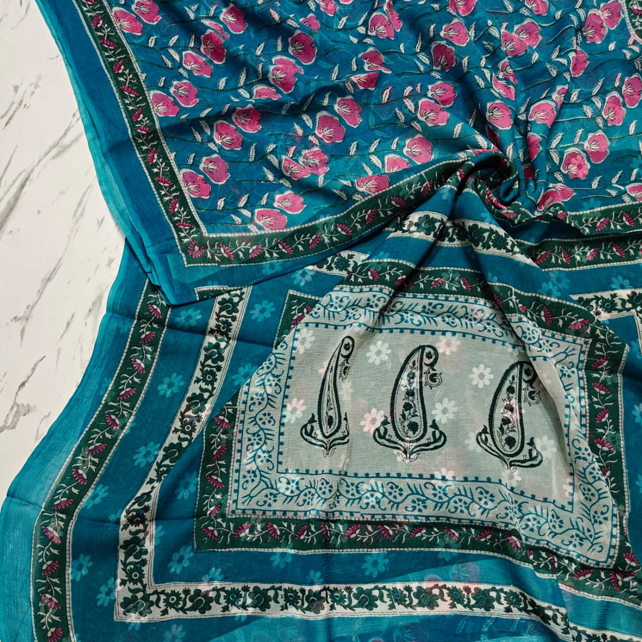 A teal blue chiffon saree with pink floral prints, a light blue-grey central panel with three black paisley motifs, and a decorative border, laid out on a white marble surface.