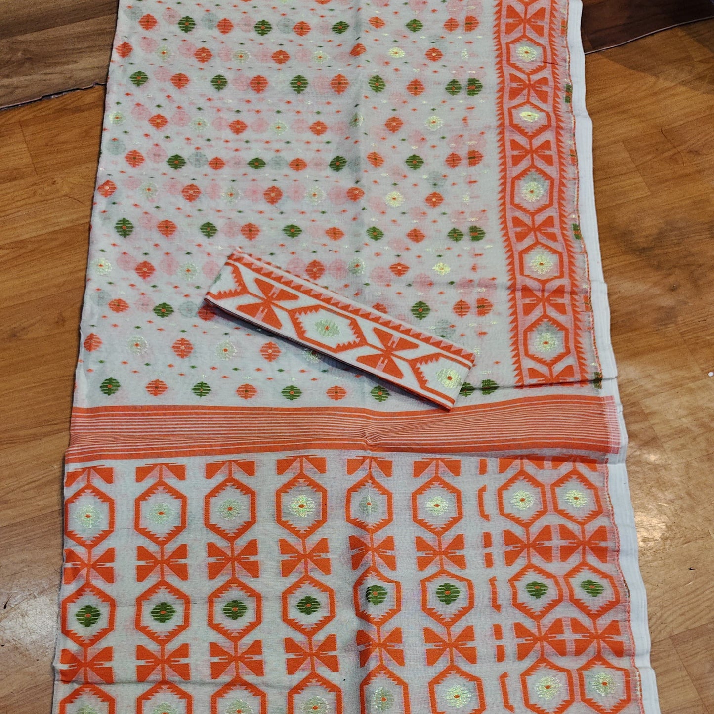 A white Banarasi silk saree with orange and green geometric patterns, laid flat on a wooden floor. The saree features a wide orange border with intricate motifs and a matching patterned blouse piece.