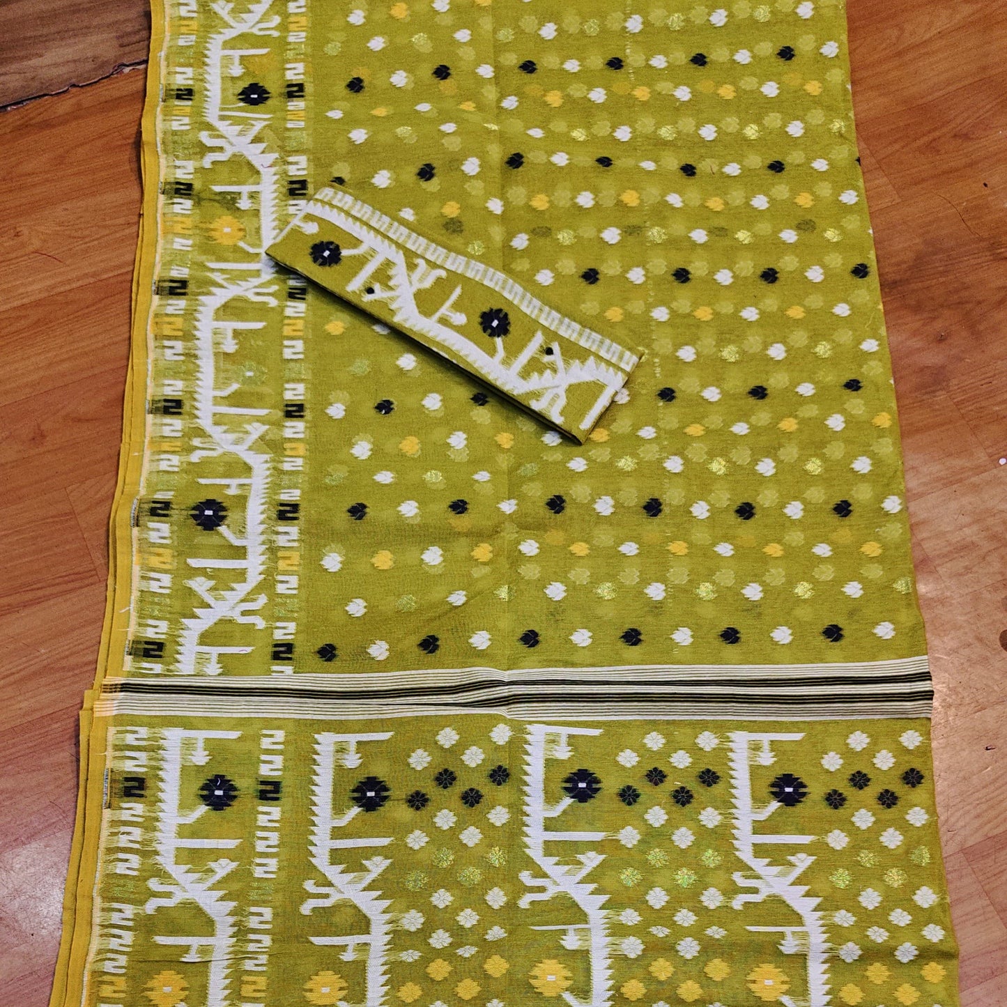 A flat-laid olive green cotton fabric set, likely a salwar suit material, featuring a printed body with white and black dots, vertical white ikat-style borders, and a matching printed fabric piece laid diagonally on top.