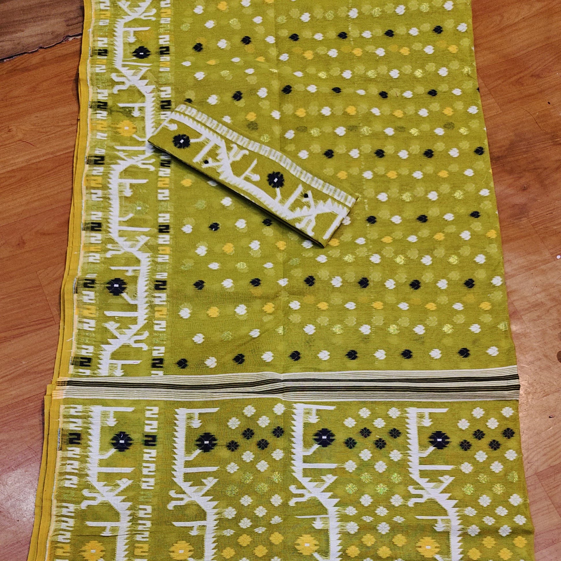 A flat-laid olive green cotton fabric set, likely a salwar suit material, featuring a printed body with white and black dots, vertical white ikat-style borders, and a matching printed fabric piece laid diagonally on top.