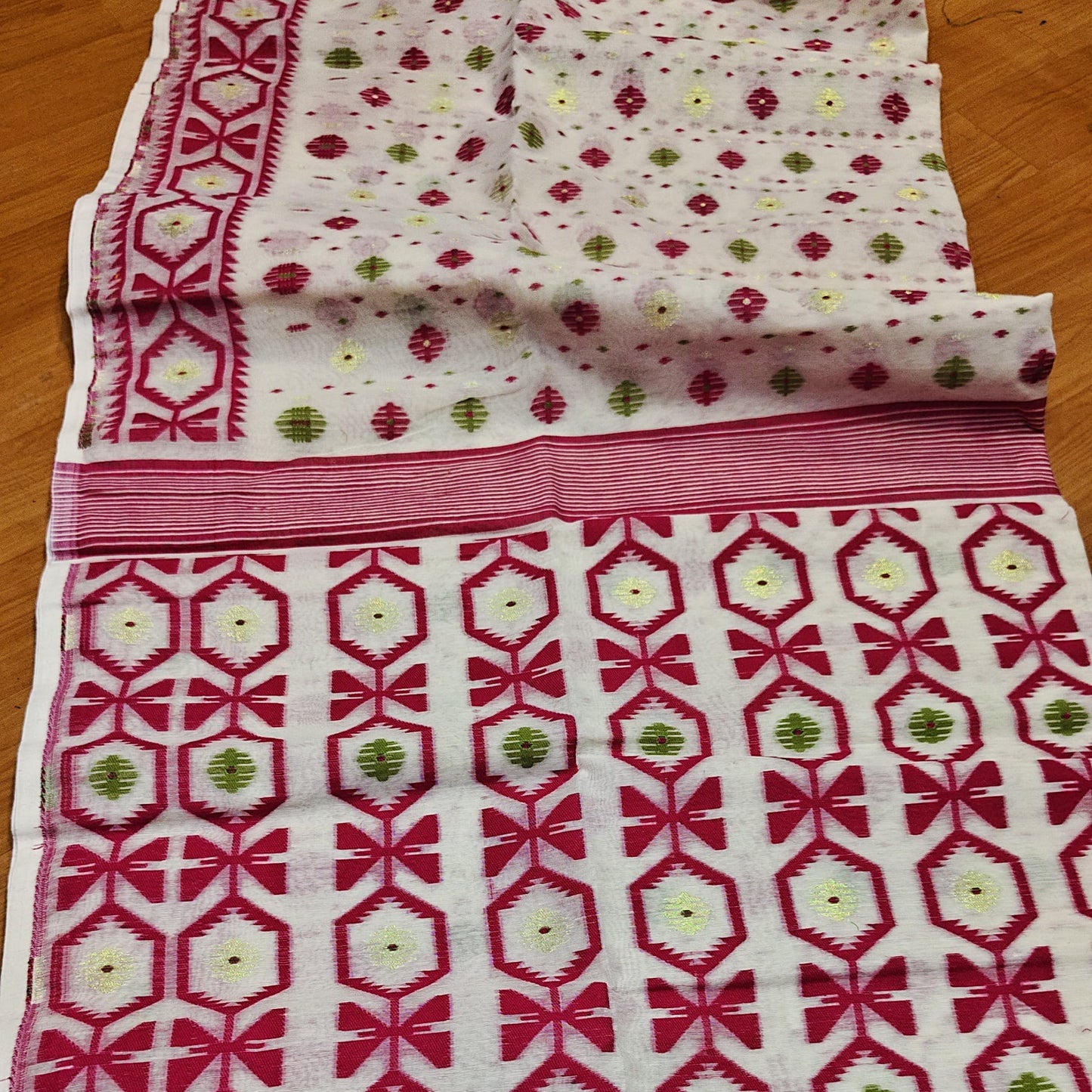 A folded and unfolded white cotton fabric, likely a saree or dupatta, featuring a vibrant magenta-pink geometric pattern with green and yellow floral accents.