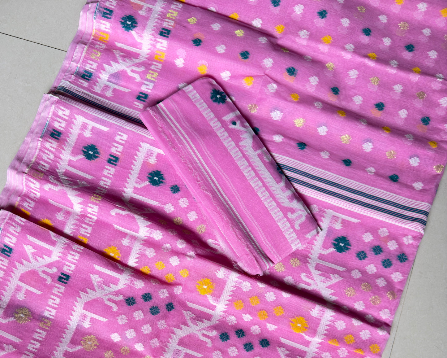 Pink cotton saree fabric with white geometric patterns, blue and yellow floral accents, and a matching pink fabric piece laid on top, photographed on a light floor.
